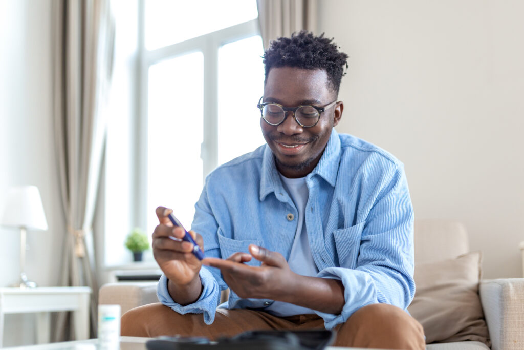 african man is sitting at the sofa at the home and taking blood from his finger due to diabetes. the daily life of a man of african american ethnicity person with a chronic illness who is using glucose tester.